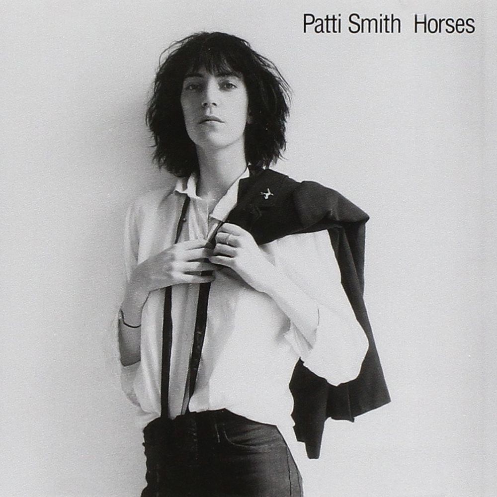 Patti Smith stands against a blank wall with a black jacket slung over her shoulder