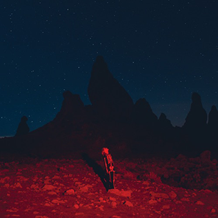 Alt text: Punisher’s album cover depicts Phoebe Bridgers standing alone in the middle of the desert at night. The desert ground is covered in rocks and lit by bright red lighting. The rest of the landscape is dominated by dark mountain rocks and a dark blue night sky. Phoebe appears very small among the rocks at the bottom-center of the cover: she’s standing straight, and you can see her from her right profile looking at the sky while her shadow is reflected on the ground.
