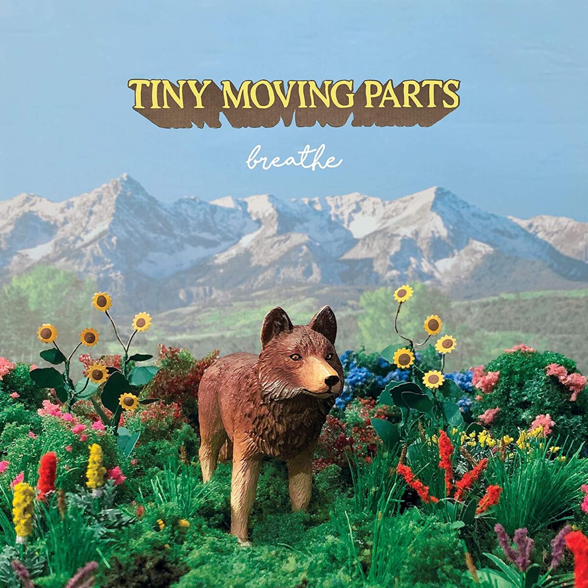 "breathe"'s album cover depicts the painted statue of a wolf standing in the middle of nature, surrounded by red and yellow flowers. There are mountains and a big blue sky behind it and "Tiny Moving Parts" is in yellow with brown outlines, in all caps.