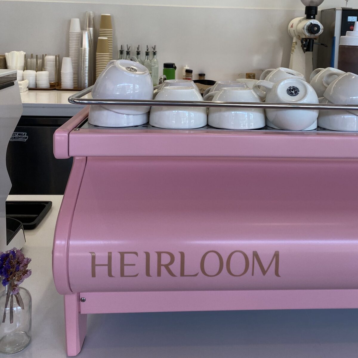 This photograph features a pink coffee machine that says "HEIRLOOM" in all caps, with white mugs sitting on it.