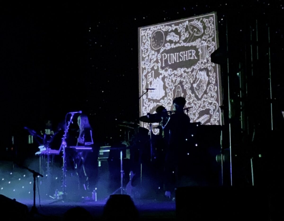Phoebe is on stage with her trumpetist. They're both wearing skeleton suits. A bright, blue light and smoke surround them. Behind them, a screen displays a black and white book cover that reads "Punisher", with scary drawings decorating it.