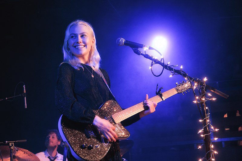 Phoebe bridgers with a purple background, performing, with a guitar in hand and smiling.