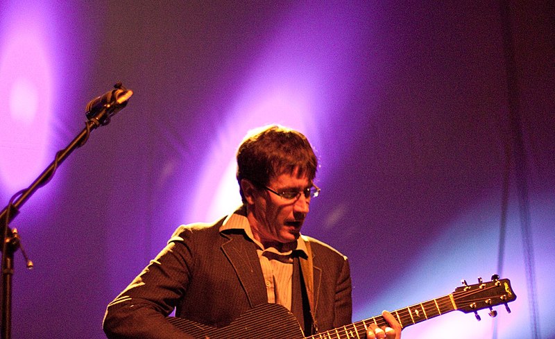 John Darnielle singing into a microphone on stage while also playing the guitar.