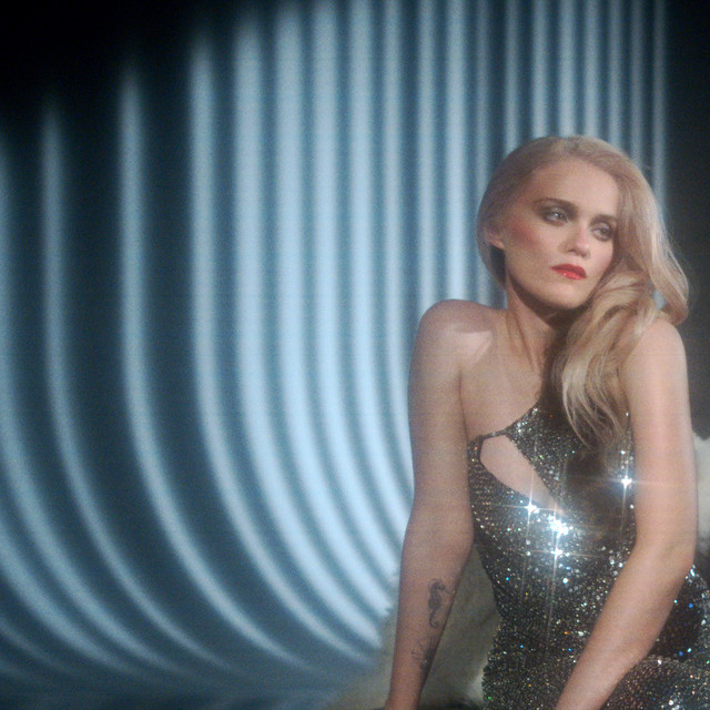 Singer Sky Ferreira sitting in front of a blue background. She is staring off into the distance and wearing a sparkly silver dress.