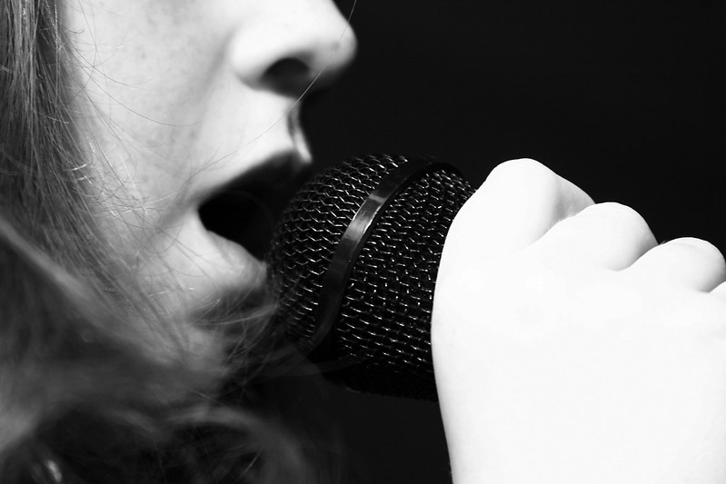 Person with pale skin holding a microphone to their lips and singing. Image in black and white.