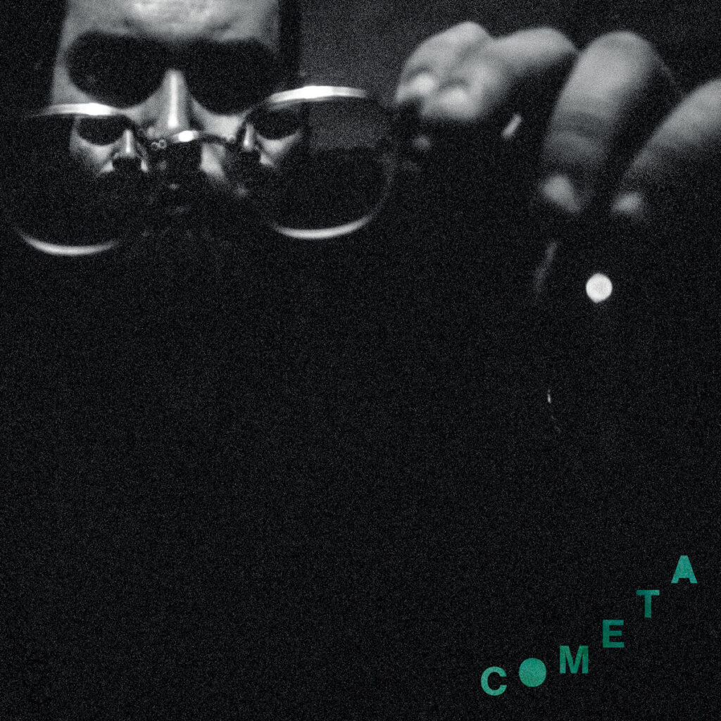Cover for "Cometa" by Nick Hakim. Features a dark close up of a man examining his glasses. Two reflections of the man can be seen through the lenses of the glasses. 