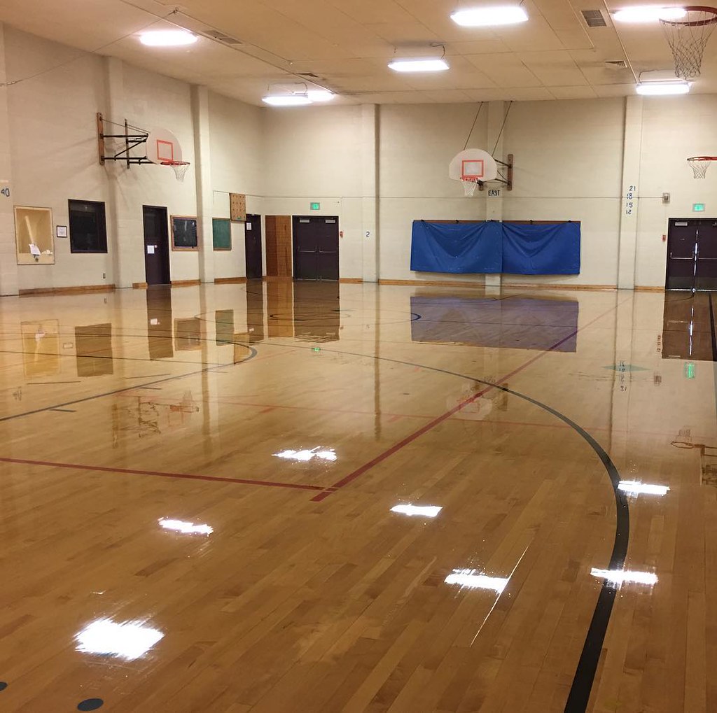 An elementary school gym floor. There are two basketball hoops, a shiny floor, and a drop ceiling with light reflecting off the floor.