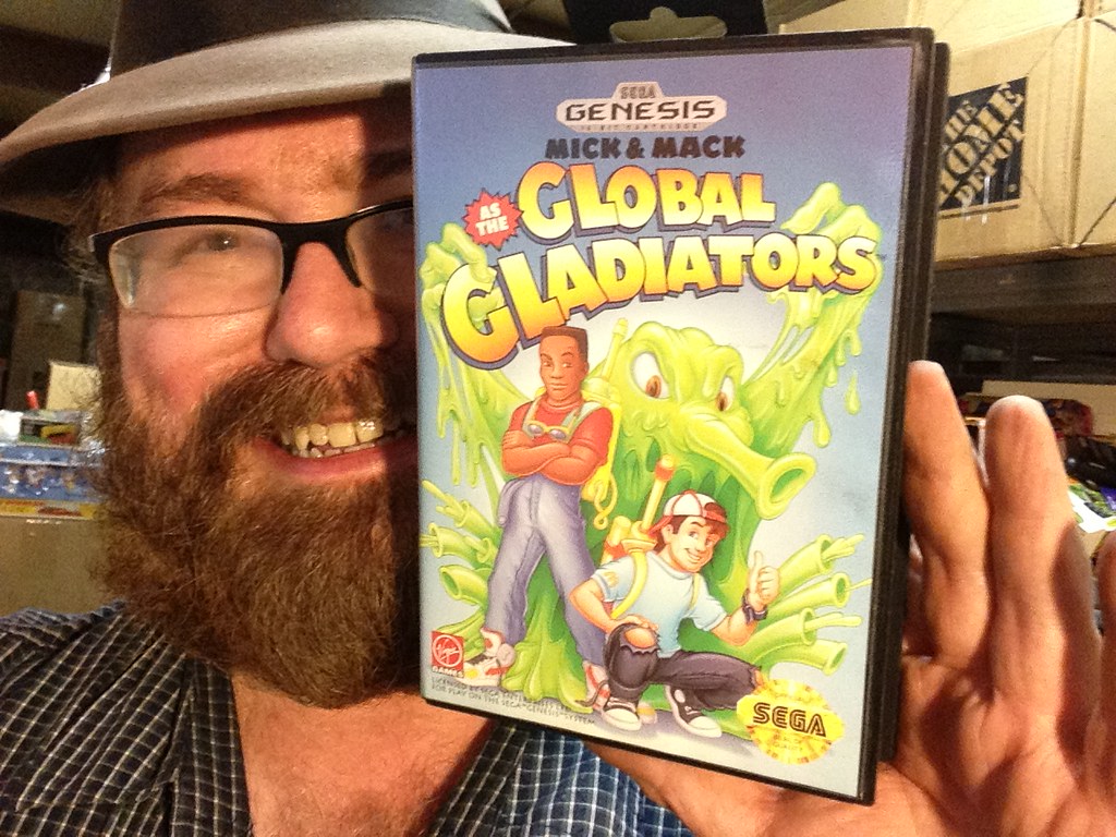 Man with a beard, glasses, and hat holds up a copy of Mick and Mack Global Gladiators for the Sega Genesis. On the cover, there is a cartoon of a big green goop monster in the background with two adolescents standing in front of it. The title of the game is in big letters at the top of the box. 