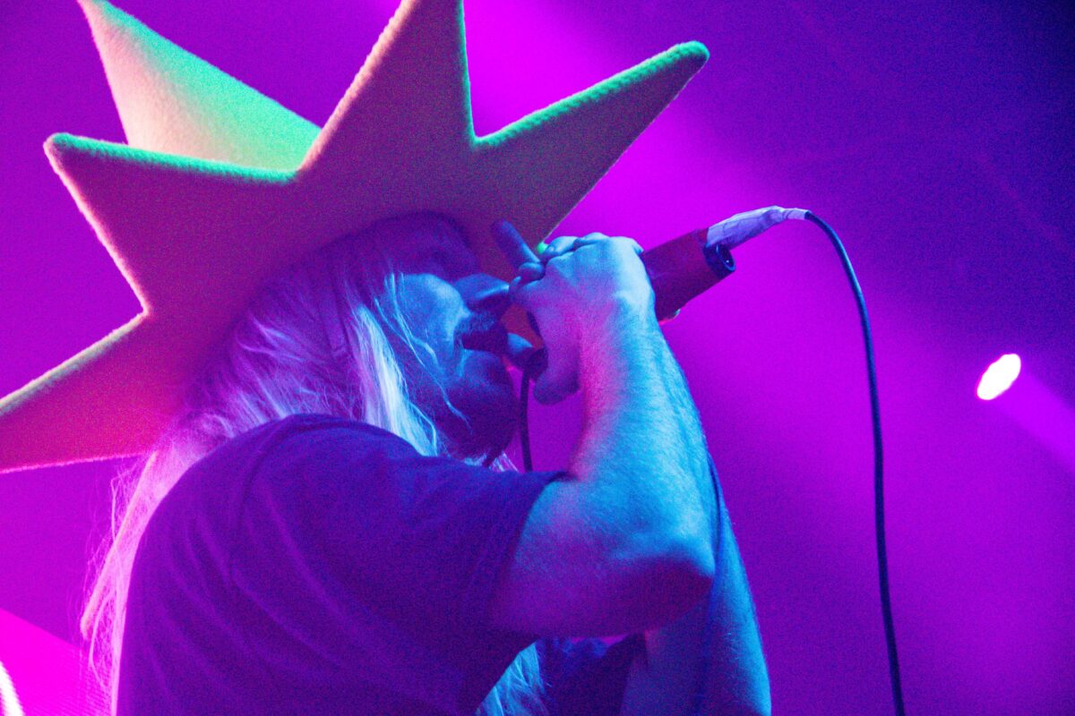 Dylan Brady of 100 gecs singing into a mic, wearing his yellow traffic-cone/wizard hat.