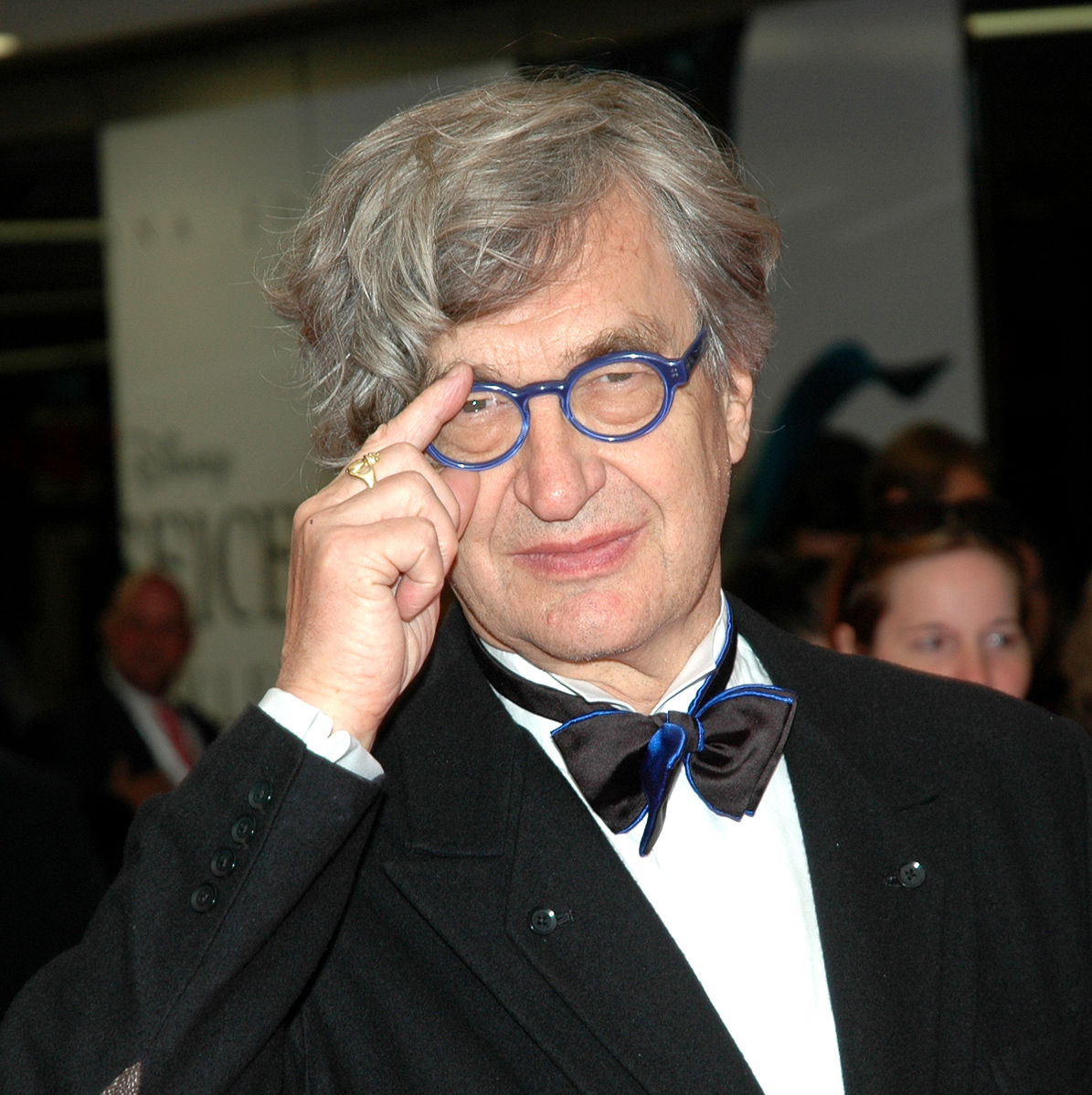 Director Wim Wenders dressed in blue glasses, a black suit, and a bow tie with blue accents. "Wim Wenders 0566" by Harald Bischoff is licensed under CC BY-SA 3.0.