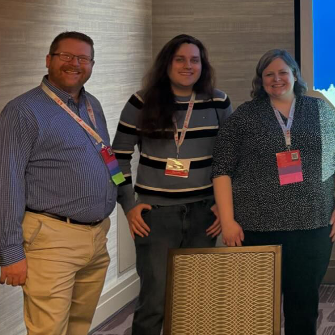 Three people standing and smiling. They have convention badges around their necks.