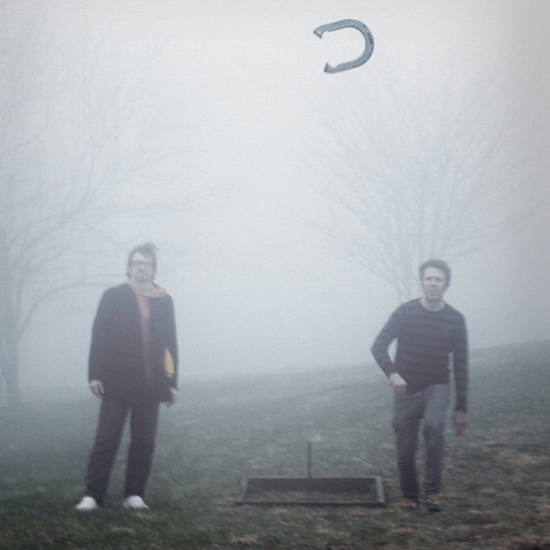 Joshua Carpenter and Finn Cohen on a foggy lawn with a horseshoe mid-air and bare trees in the background.