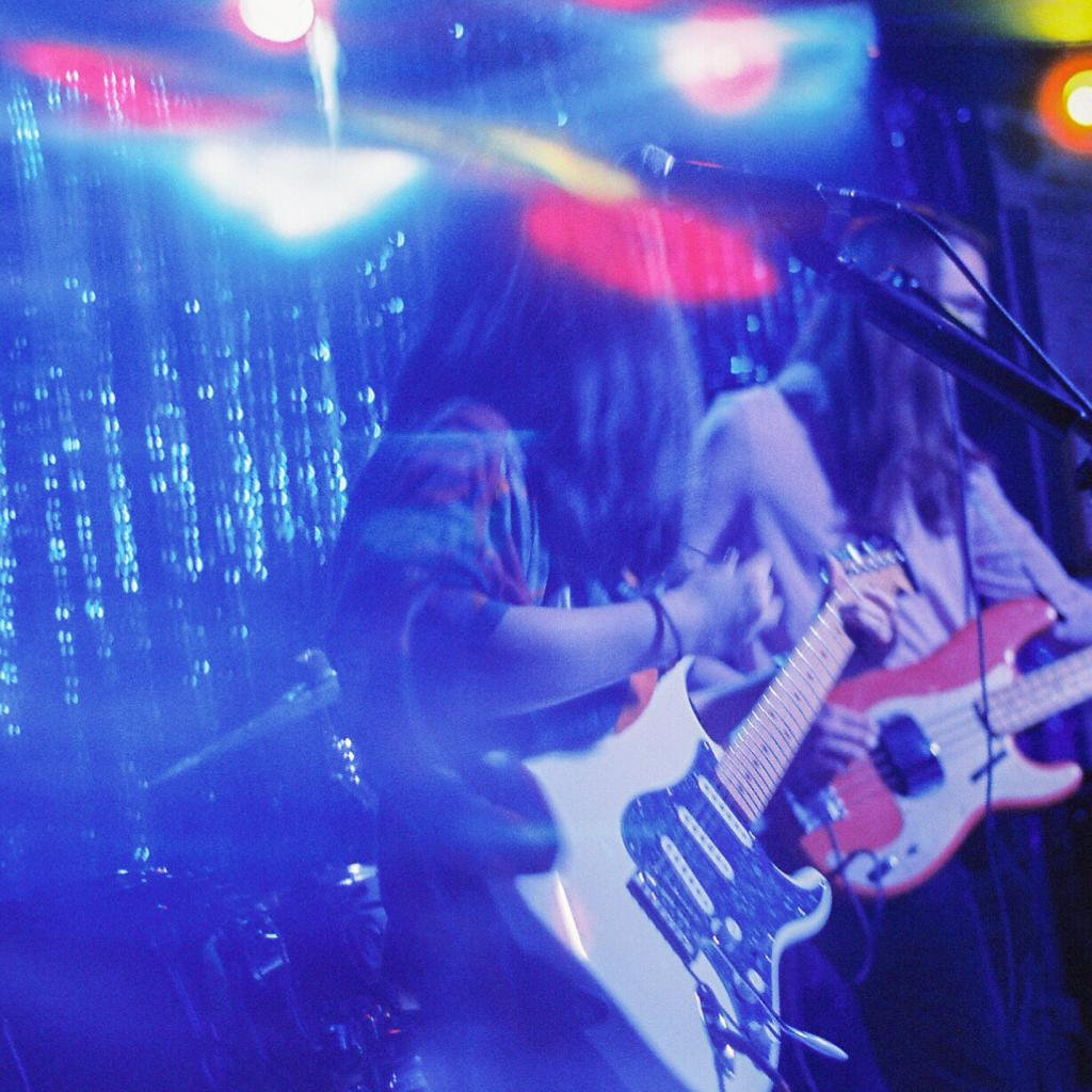 Musicians from My Sister Maura playing guitars on a stage with vibrant lighting in blue and red hues.