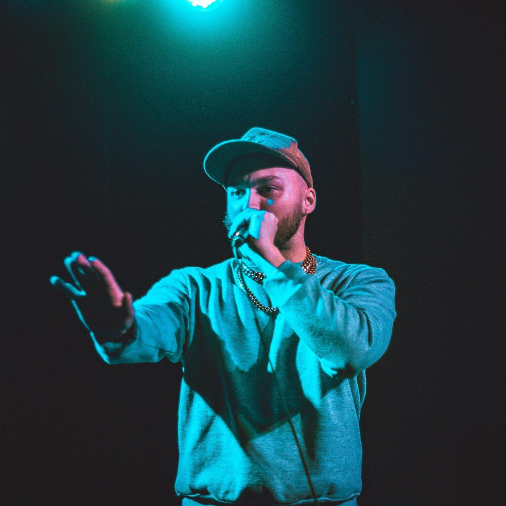 Sk the Novelist performing on stage with a microphone under blue-green lighting.