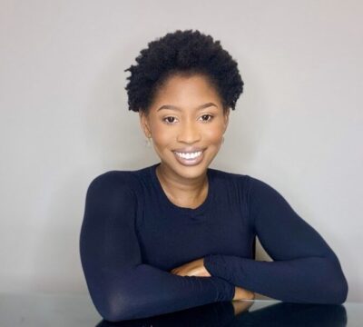 A smiling person with short curly hair, wearing a dark long-sleeved top, sits with arms crossed on a table.
