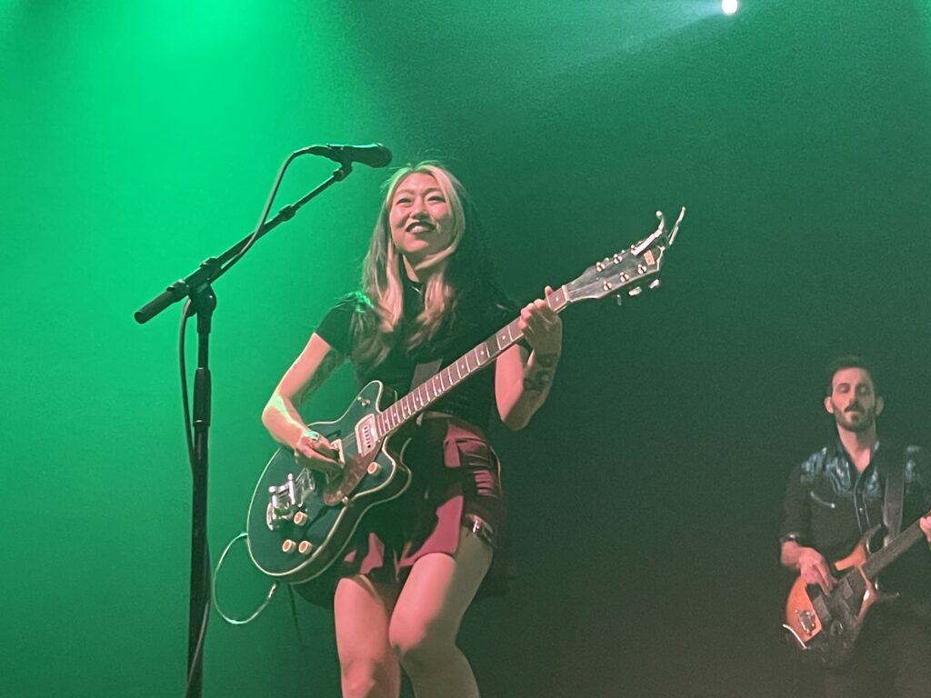 The Crane Wives singer, Emilee Petersmark playing guitar while backlit by green light. Bassist Ben Zito can be seen in the background.