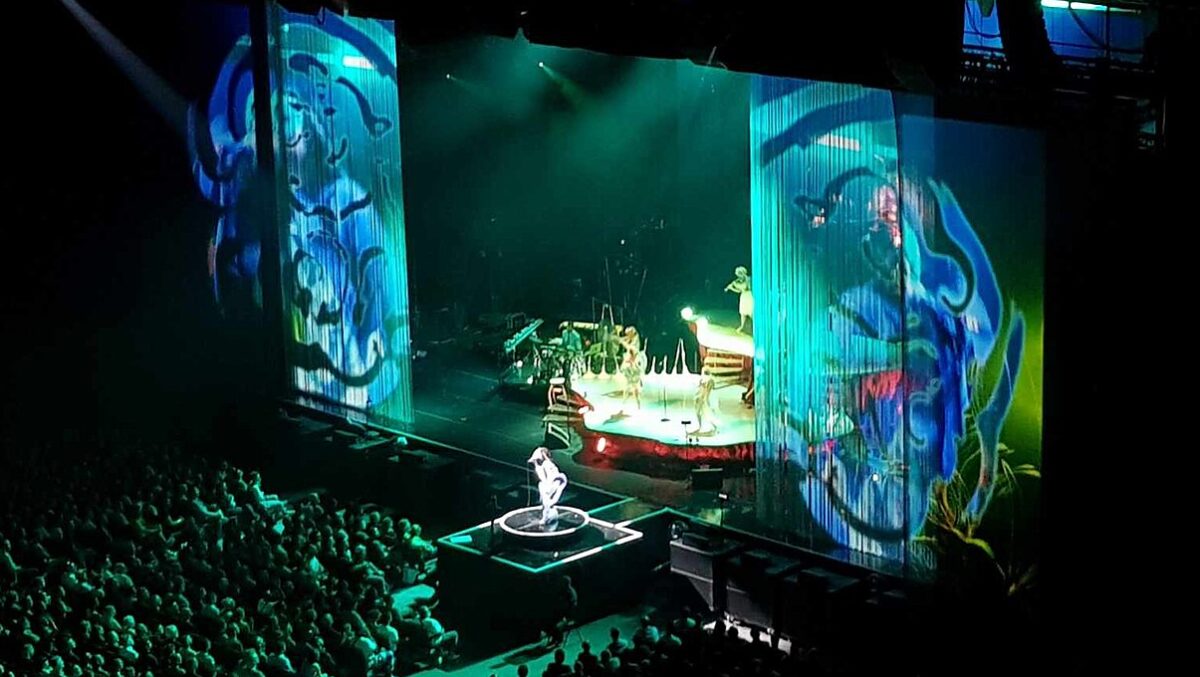 Björk performing on a stage lit in blue and green in front of people playing instruments.