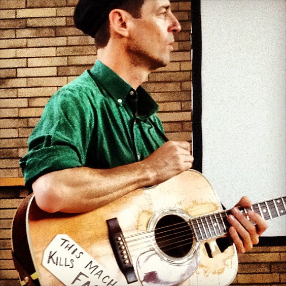 Woody Guthrie stands with guitar, facing away from the camera