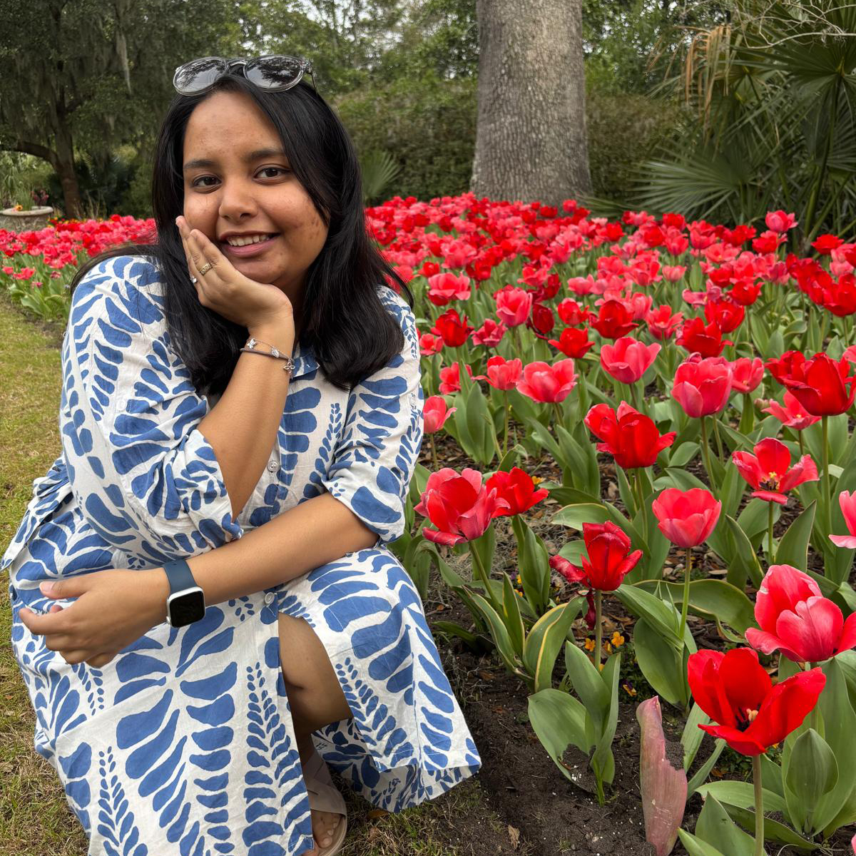 Woman kneeling beside a bed of red tulips in a garden wearing a blue and white patterned dress.