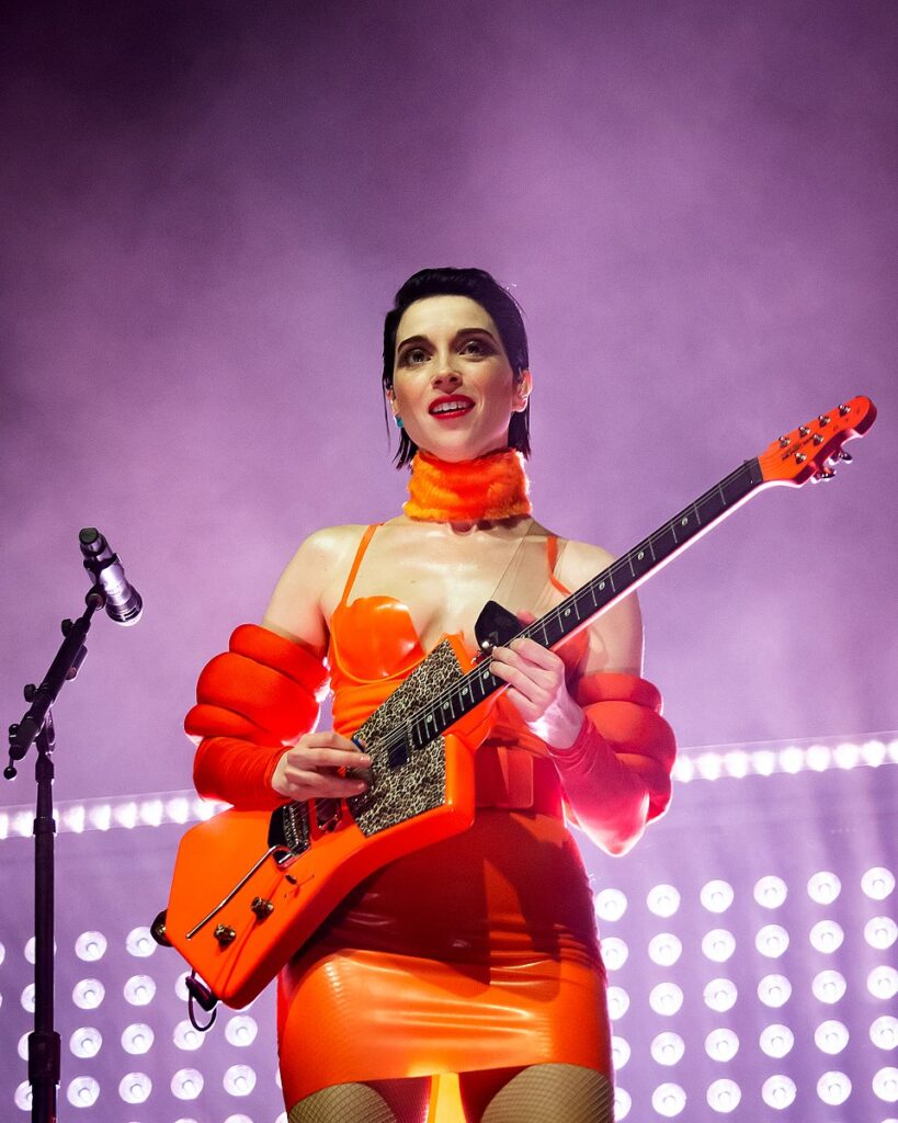 St. Vincent performing with a guitar in an orange outfit.