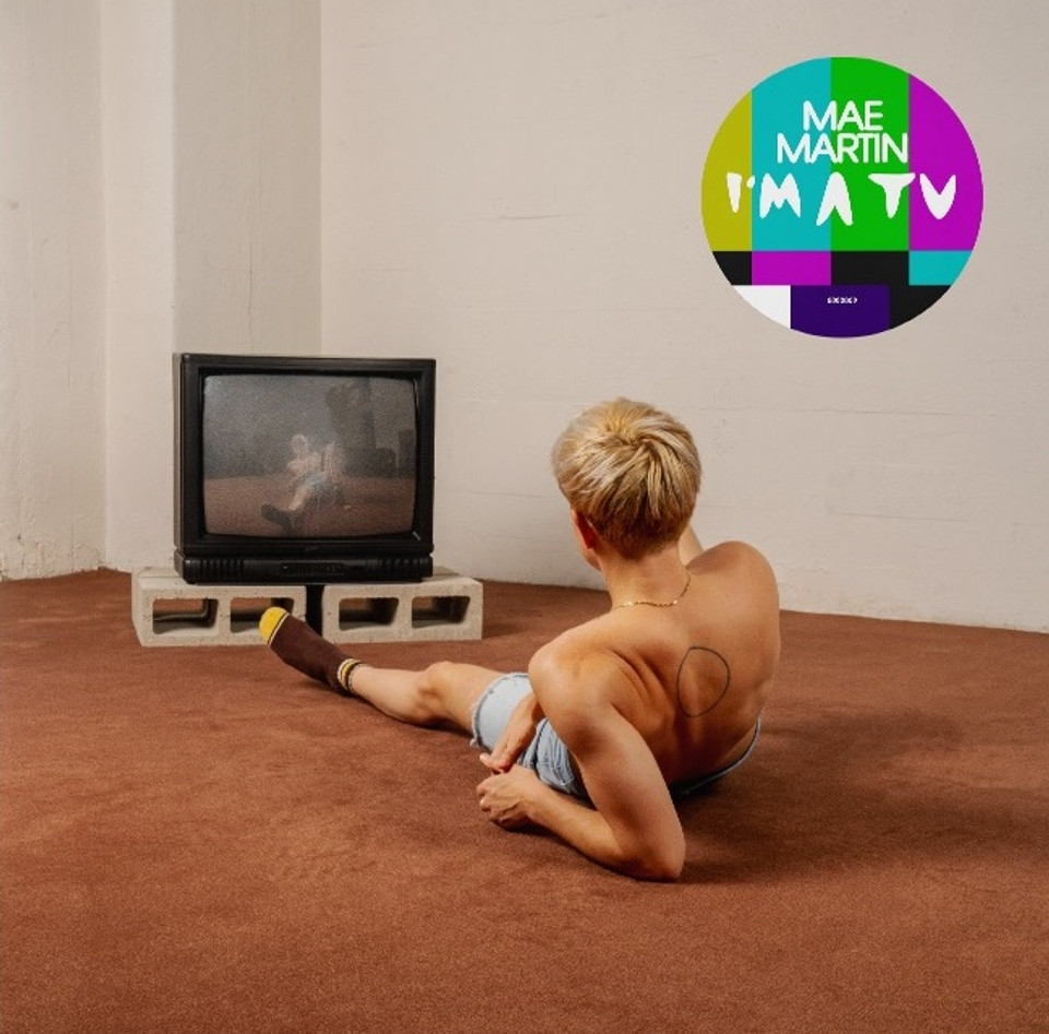 Mae Martin's "I'm A TV" album cover. A person with short hair sitting on a brown carpet watches an old TV on concrete blocks. A colorful emblem is on the wall.