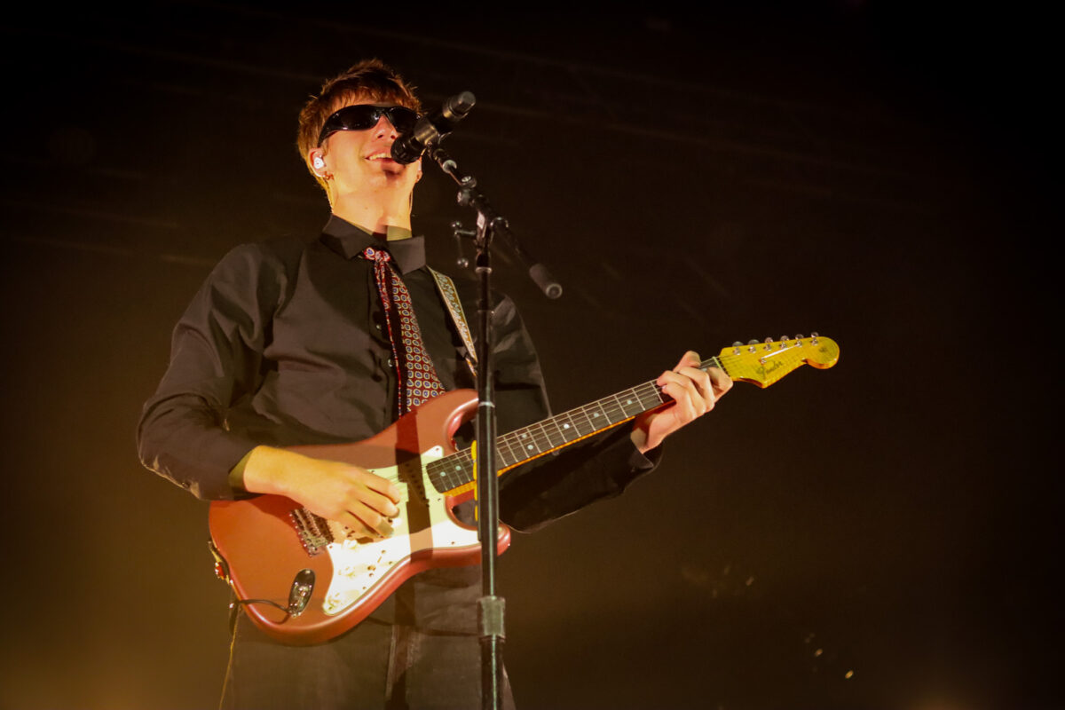 Malcolm Todd stands at microphone stand smiling with his electric guitar.