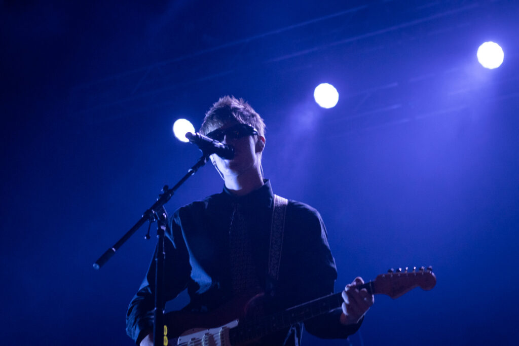 Malcolm Todd is singing into microphone with his guitar and blue light behind him.