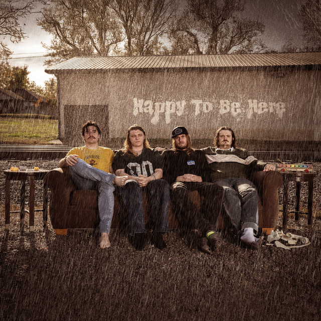 4 men sitting on a couch outside in the rain with "happy to be here" written behind them