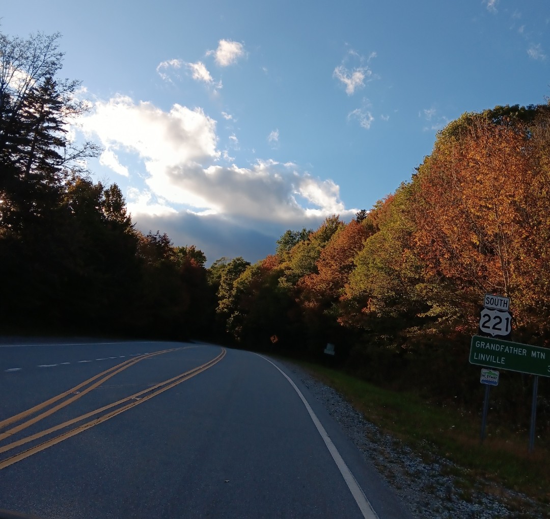 Stretch of road with red and orange leaved trees on the side and a highway sign indicating Grandfather Mountain and Linville