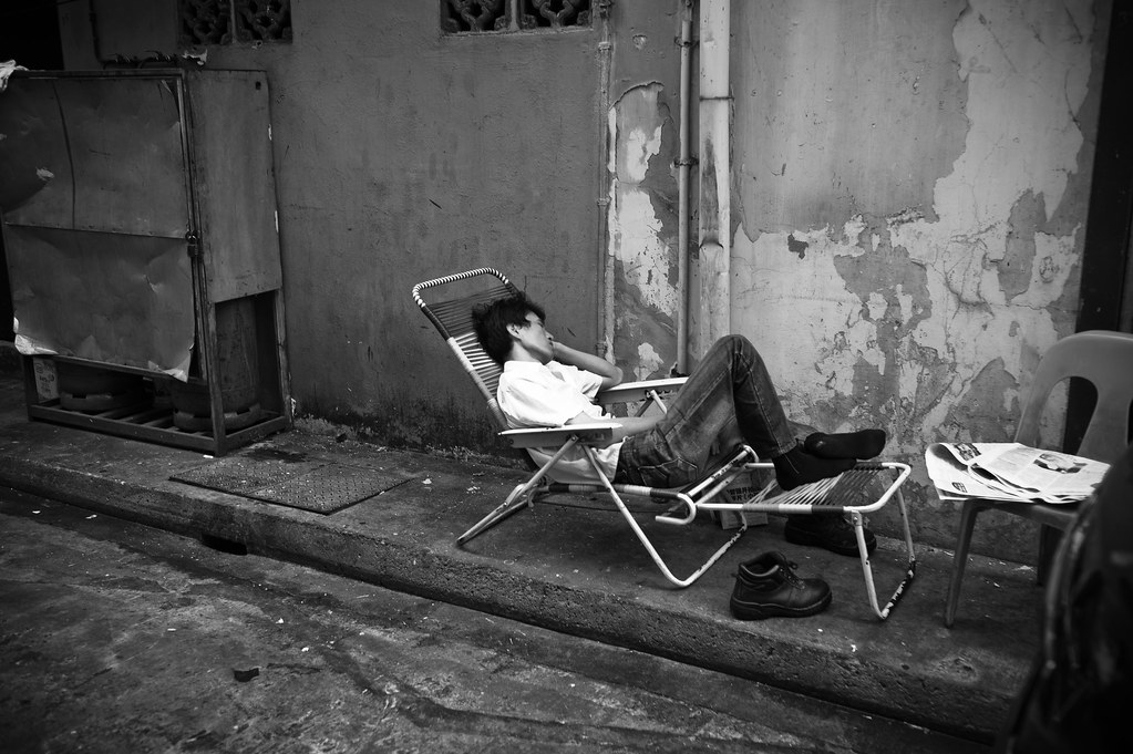 Black and white image of a man asleep on a lawn chair on the sidewalk in a city