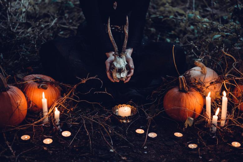 Dark scene with hands holding a deer skull in the center over a candle with and pumpkins and candles on either side of the skull