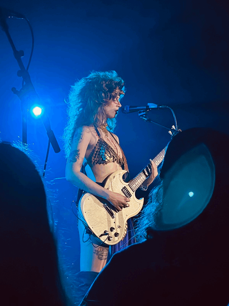 The Rat Queen Performing a Guitar Solo A woman with a eighties blow-out a scale-mail bikini shining under blue stage lights as she plays her guitar. Photo by Vernice Enierga, used by permission.