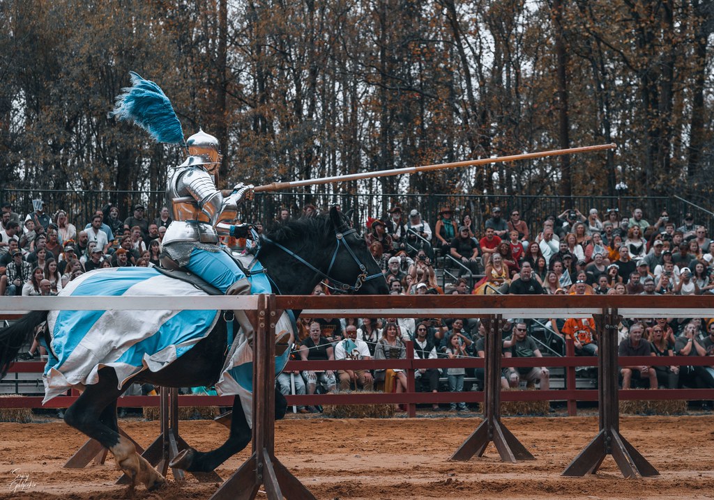 A person in armor and blue accents on top of a horse holding a lance before an audience.  