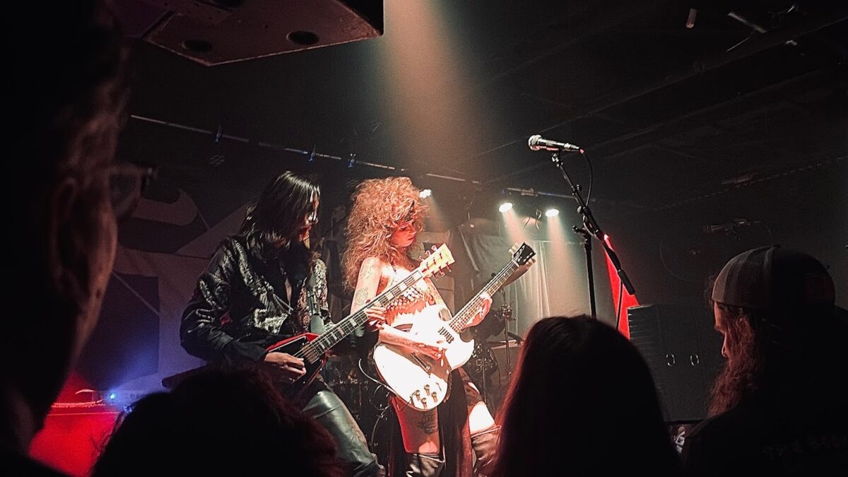 A woman with an eighties blow-out in a scale mail bikini strumming a white electric guitar next to a man with long black hair in a black leather tailcoat strumming a red electric guitar. Photo by Vernice Enierga, used by permission.