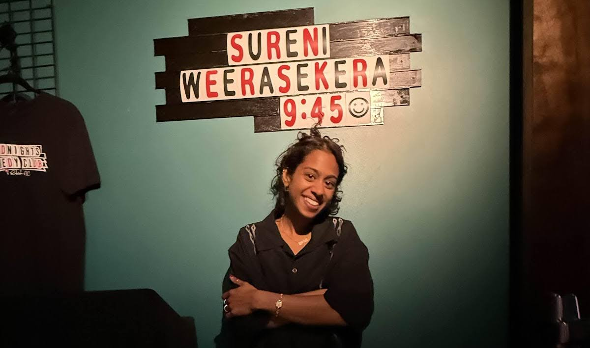 A smiling woman in an oversized black dress shirt with an updo standing in front of a sign that reads "Sureni Weerasekera 9:45." Photo by Killian Le, used by permission.