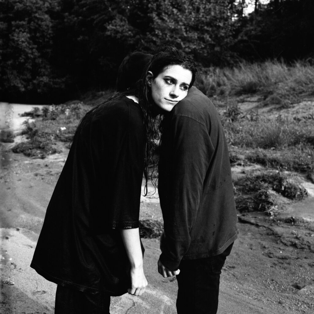 Two people from the band Monsoon standing close on a sandy outdoor area, with one resting their head on the other's shoulder, in a black-and-white photograph.