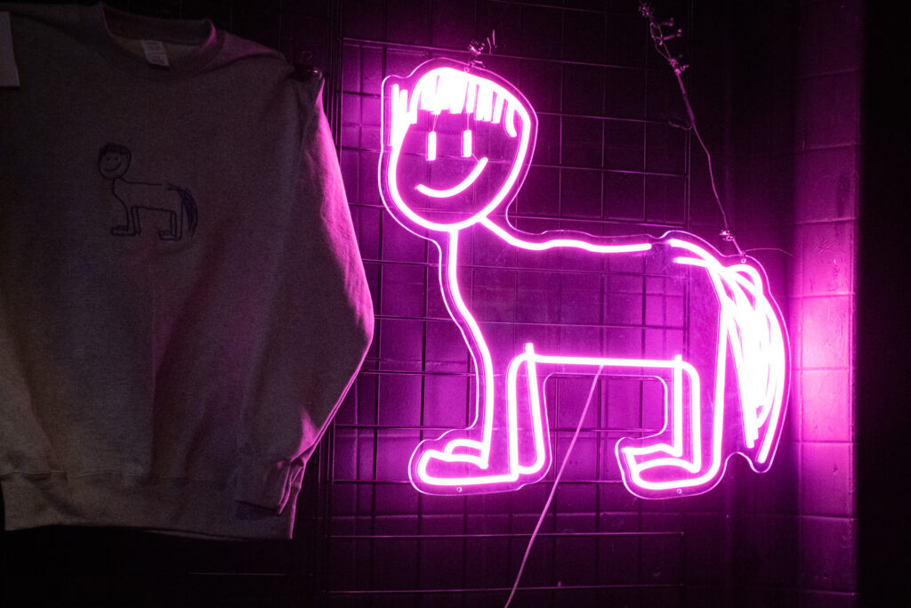 Pink neon sign of horse with bowl cut next to gray crewneck that has horse with bowl cut image on it.