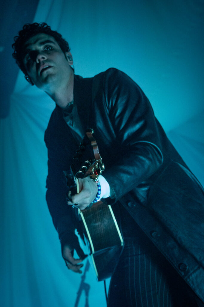 Del Water Gap in black trench coat playing acoustic guitar with blue lights.