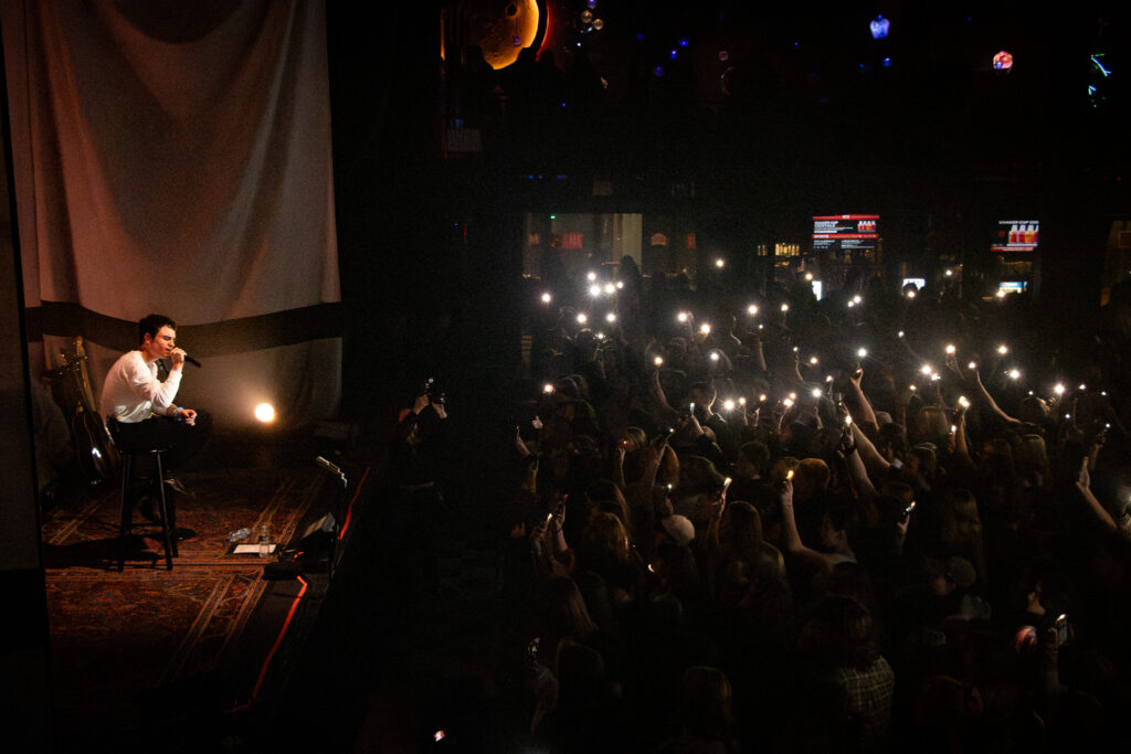 Del Water Gap sitting on stool singing on stage with audience holding up flashlights.