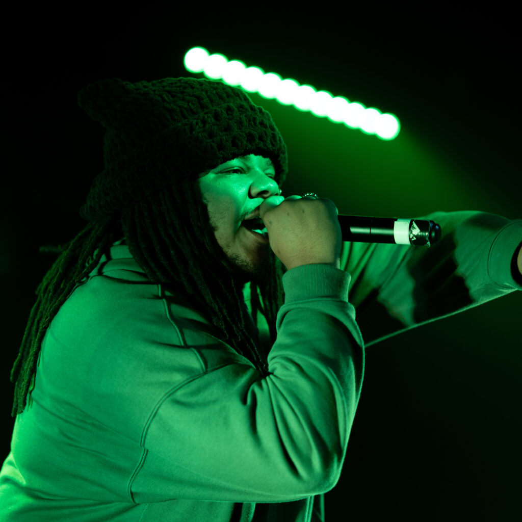 Person wearing a beanie and hoodie performing on stage with green lighting.