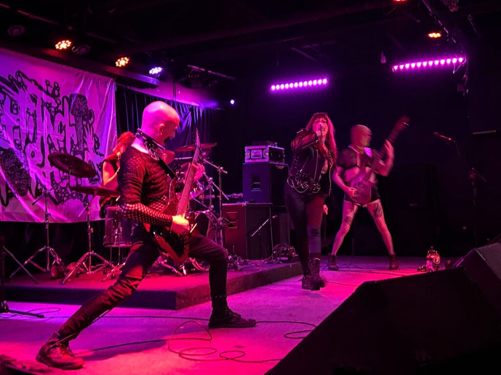 A pink-red lit stage with three members of Hitachi Torture onstage playing. By Killian Le, used by permission.