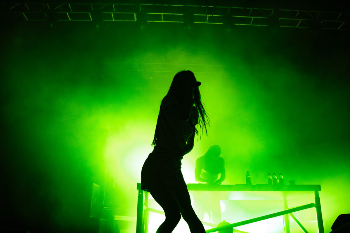Snow Strippers in green lighting on stage. Tatiana Schwainger silhouette with Graham Perez DJing behind the table.