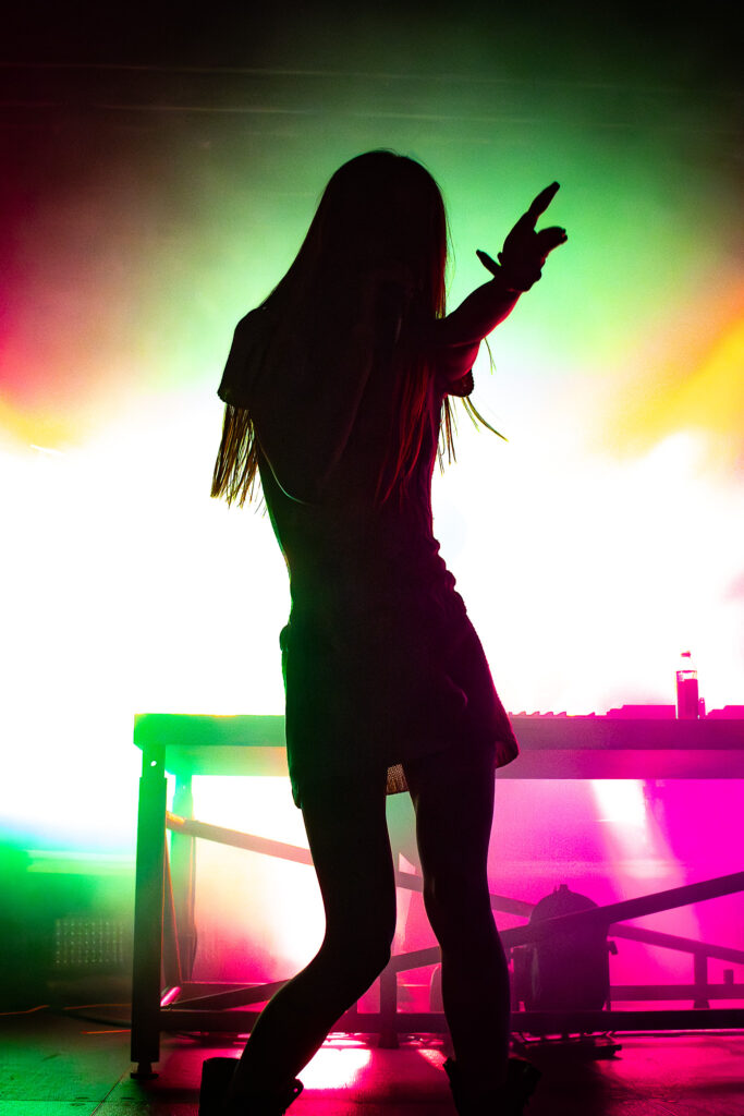 Snow Strippers' Tatiana Schwainger pointing finger gun with colorful lights behind.