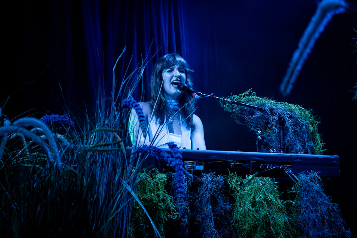 Madison Cunningham sits at piano with moss and reed plants surrounding her. She is singing into a microphone in blue lighting.