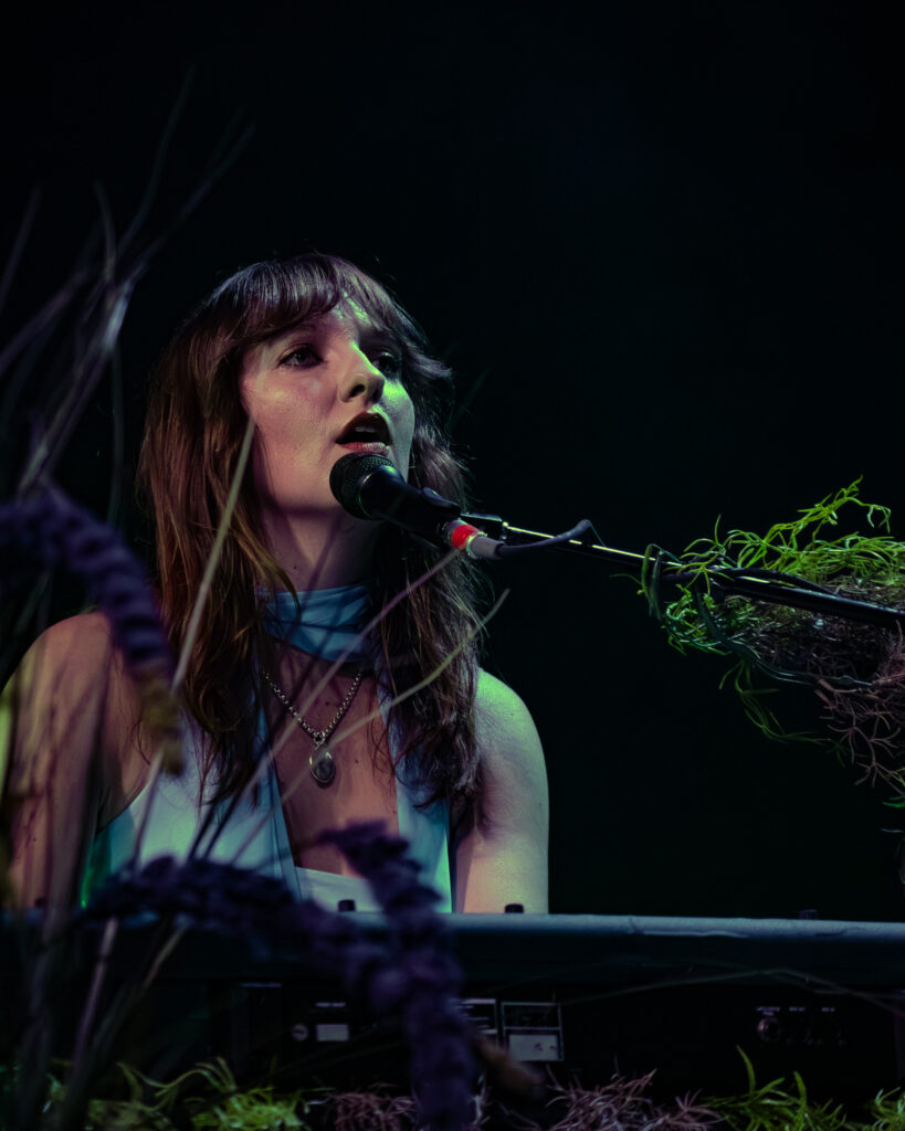 Madison Cunningham sitting at the piano through the reed plants looking across stage.
