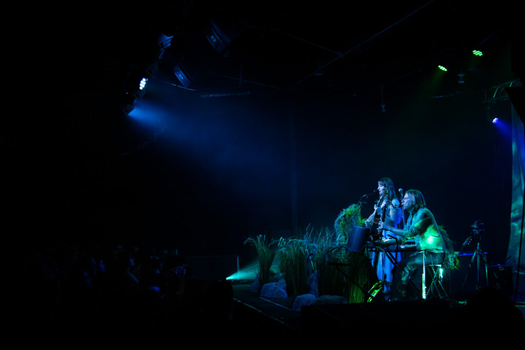 Wide shot of Madison Cunningham playing guitar and singing while Jesse Chandler plays bass clarinet on stage. The crowd is watching in blue lighting.