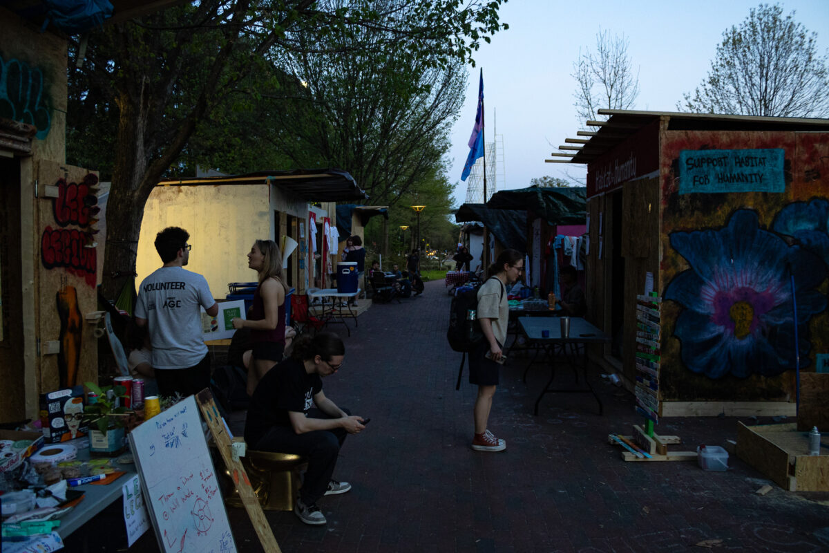 A few students standing between fully built shacks during Shackathon, the walls of the shacks painted with many tables and people in front of them