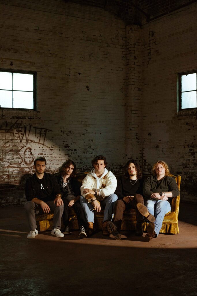 5 men sitting on a brown corduroy couch in a graffitied, exposed brick building