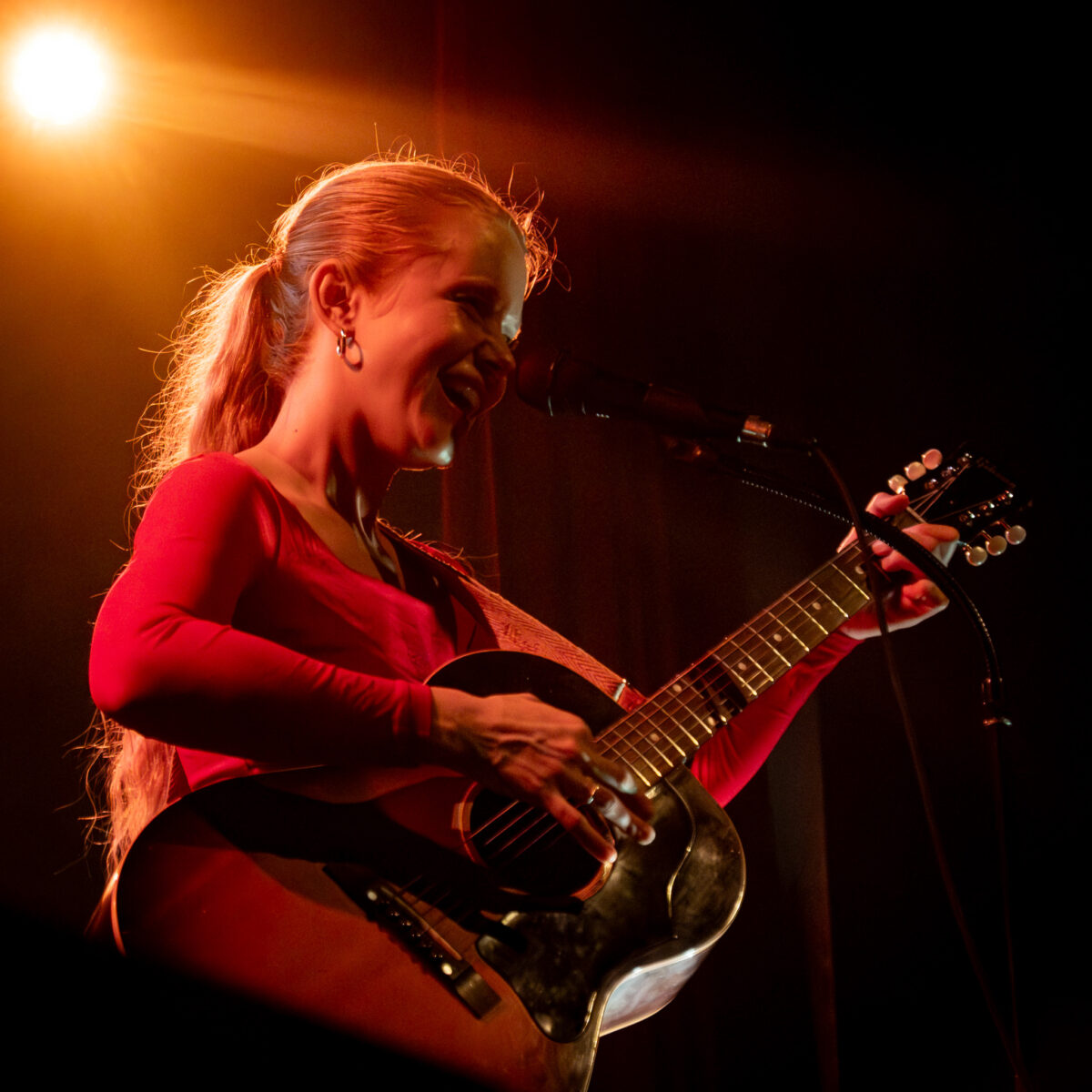 Alice Phoebe Lou smiling at microphone stand with acoustic guitar in hand. Warm light shines from above.