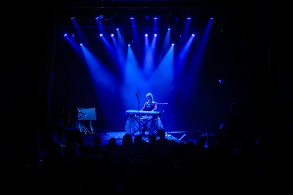 Alice Phoebe Lou sitting center stage at keyboard with many blue spotlights on her.
